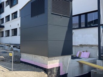 Großes graues Lüftungsgerät zur Vorfilterung von Außenluft, platziert auf einem Betonsockel neben einem modernen weißen Gebäude mit vielen Fenstern. Das Gerät steht auf einer Baustelle, erkennbar an losem Kies und einer Baustellenabsperrung im Vordergrund. | © Walter Bösch GmbH & Co KG