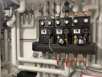 Komplexe Installation von Heizungs- und Wasserversorgungsrohren in einem technischen Raum. Das System beinhaltet mehrere weiße und schwarze Rohrleitungen, Ventile, Manometer und Pumpen, die organisiert an einer Wand montiert sind. | © Walter Bösch GmbH & Co KG