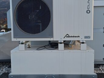 Große, industrielle Klimaanlage der Marke Clivet mit dem Logo von bösch auf der Vorderseite, auf einem Dach installiert. Die Anlage ist grau und hat einen großen, kreisförmigen Lüfter auf der linken Seite. Im Hintergrund sind Berge und ein leicht bewölkter Himmel sichtbar.