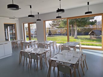 Heller Speisesaal mit großen Fenstern, die einen Blick auf eine gepflegte Grünfläche bieten. Der Raum ist mit mehreren weißen Tischen und grauen Stühlen ausgestattet. An der Decke hängen schwarze Pendelleuchten. | © Marius Projekt ZT GmbH Marius Projekt ZT GmbH