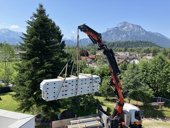 Ein roter Kran hebt ein großes, weißes Lüftungsgerät auf ein Dach. Im Hintergrund sind grüne Bäume und beeindruckende Berge unter einem klaren blauen Himmel zu sehen.